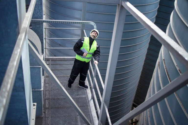 factory worker standing metal platform industrial storage tanks looking up visual inspection silos food production