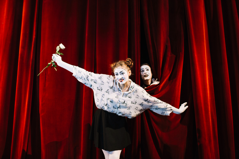 female mime holding white rose performing stage with male mime peeking curtain